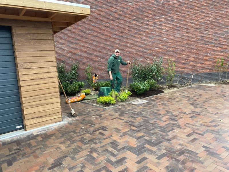Pascal van den Berg in bedrijfskleding bij een pas aangeplante border naast een houten berging, met bestrating van gebakken klinkers in keperverband